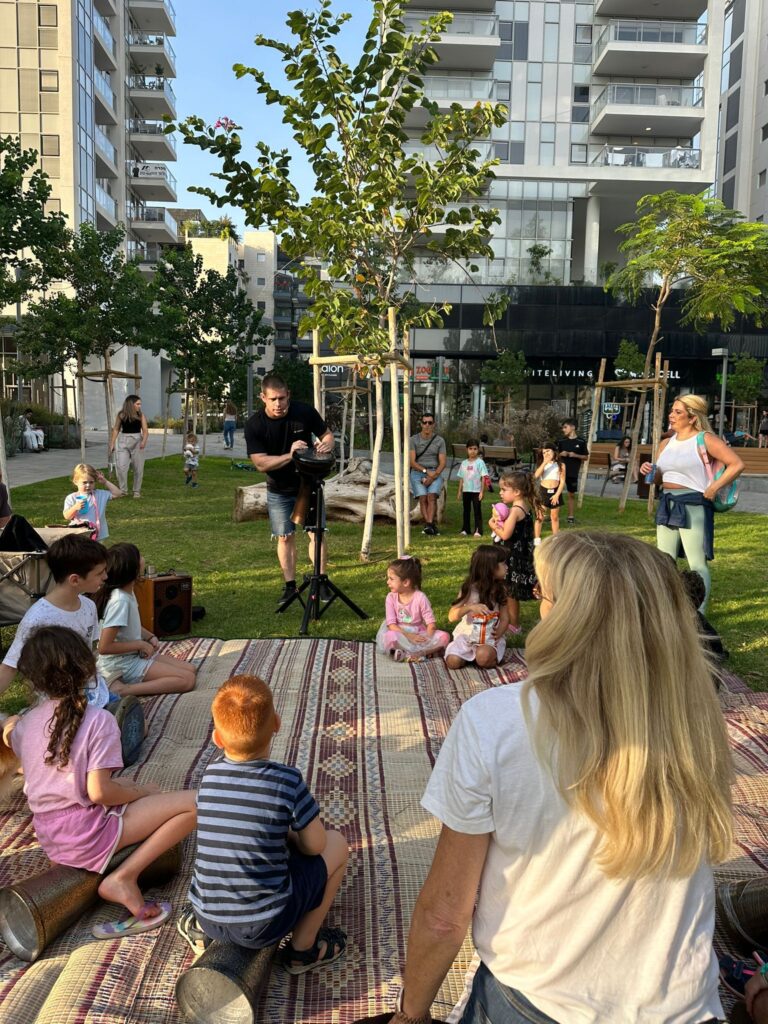 Man plays drums outdoors with group of children listening